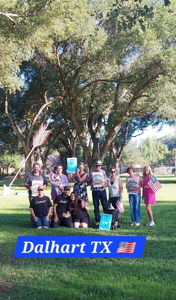Dalhart cleanup volunteers kneeling and standing on a lawn under shade trees, holding event signs and small American flags