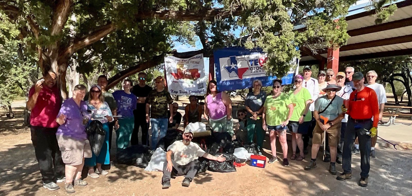 About thirty geocachers gathered under live oaks beside a park pavilion, with full trash bags at their feet and a TXGA banner hung behind them