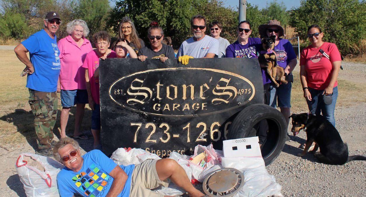 A cleanup crew posing behind a large salvaged Stone’s Garage sign and a tire they pulled out, with one volunteer lounging on the bags in front